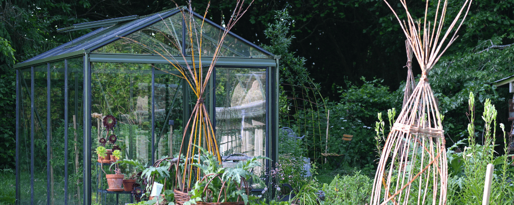 Serre Luxia LAMS vert olive de 2,36 × 3,09 m, avec des plantes en pots à l’intérieur, entourée d’un potager et de tuteurs en branches.
